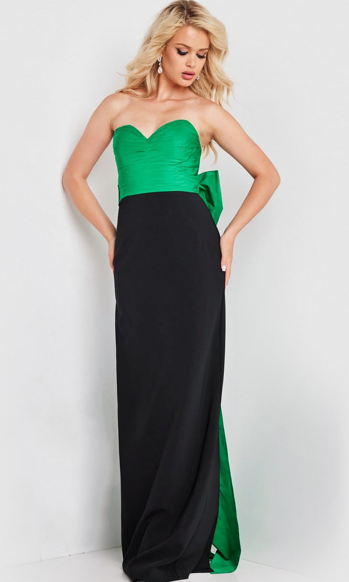 Woman wearing the Long Prom Dress: Jovani 36670, a strapless gown with a ruched green bodice, oversized green back bow, and long black skirt, stands against a plain light background.